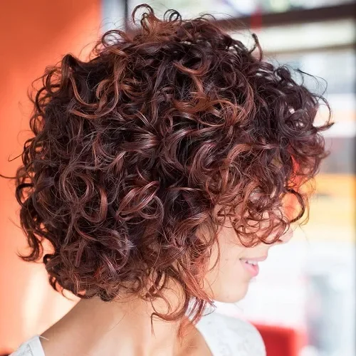 Stacked curly bob with blended bangs and dense spirals in deep mahogany brown.