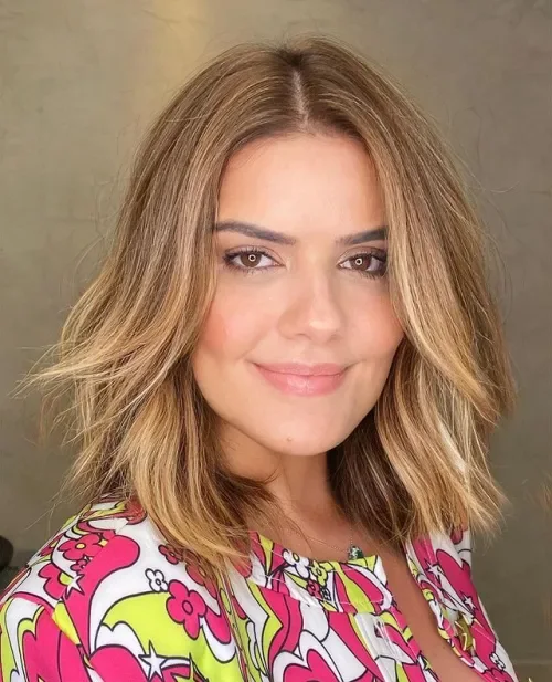 Soft layered long bob with collarbone length, face-framing pieces, warm blonde highlights, and subtle waves.